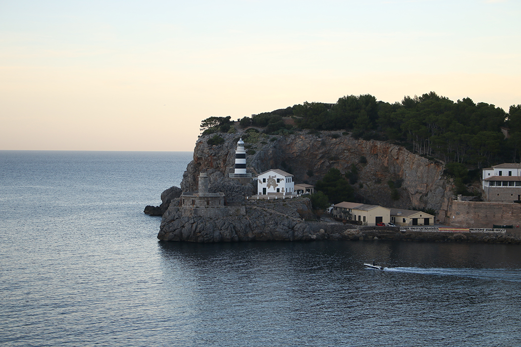 "Faro", fotografía tomada no porto de Sóller, Mallorca, o 11-12-23. Fotografía do Faro do porto de Sóller, tomada a moita distancia dende o extremo oposto da baía coa luz tenue do solpor. É un faro baixo, en raias azuis e brancas, cun pequeno edificio branco en ruínas ao carón. Vese unha parte do cabo onde está situado, mais pola esquerda albíscase o horizonte no mar aberto.