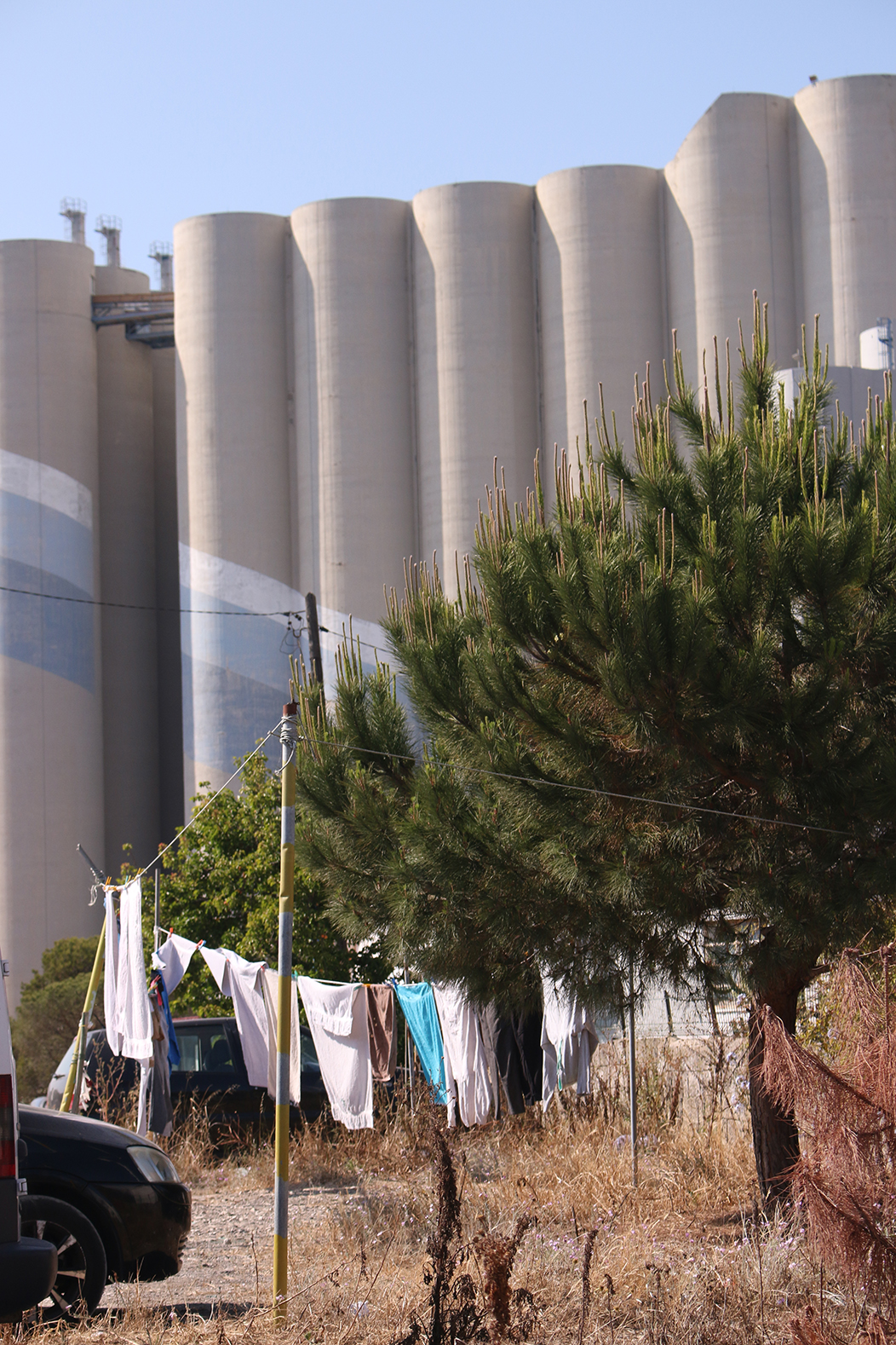 "Contraste", fotografía tomada en Trafaria, Portugal, o 2-6-23. Mostra en primeiro plano un solar utilizado pola veciñanza como aparcamento e como tendedeiro de roupa, con algunha vexetación seca e mais un pequeno piñeiro. De fondo, os silos da terminal portuaria de Silopor, unha mole industrial que coroa a paisaxe do pobo de Trafaria.