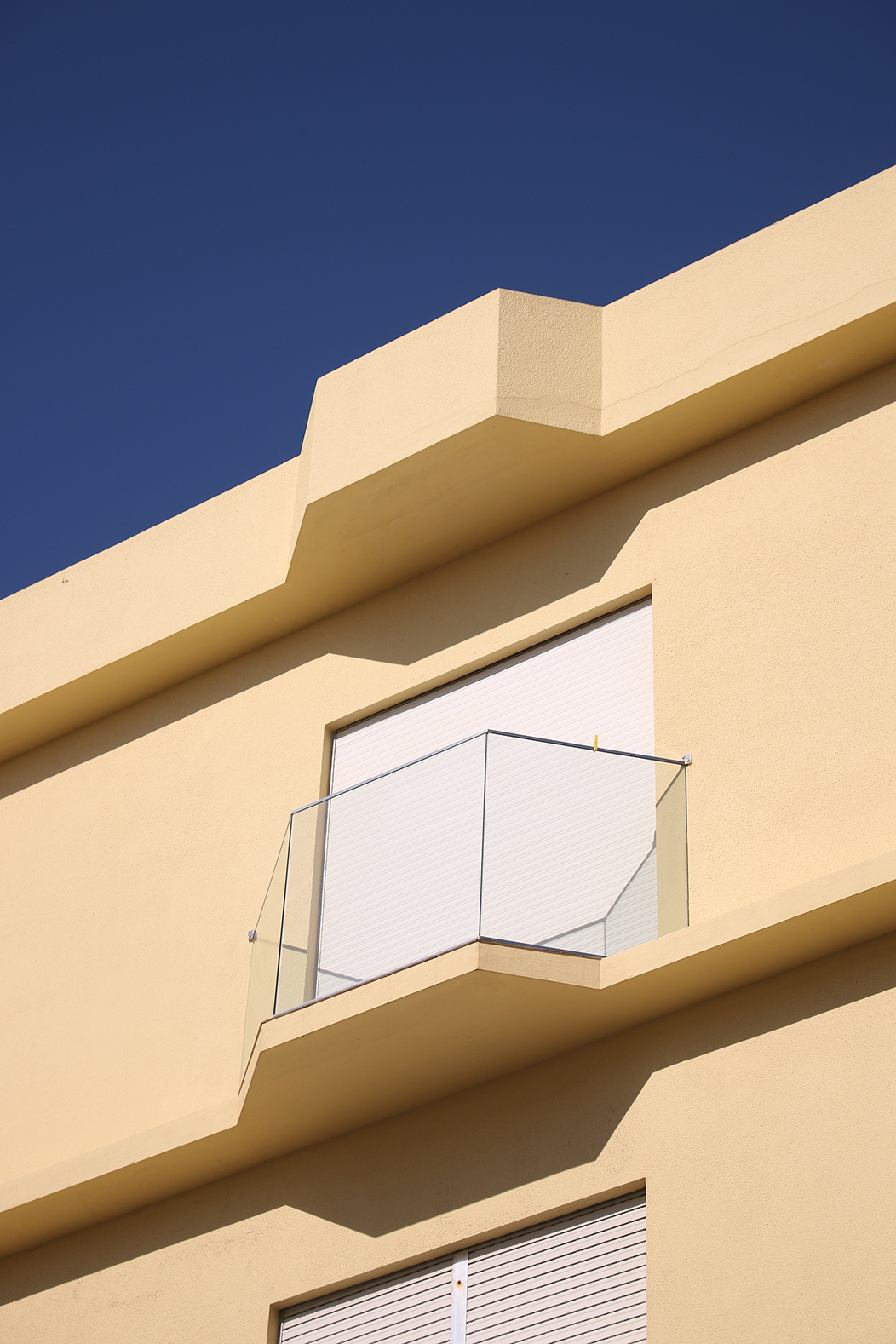 "Xeometría", fotografía tomada en Vila Praia de Âncora, Caminha, Portugal, o 4-4-23. Mostra un balcón dun edificio de Vila Praia de Âncora, con estética de vila costeira e marcada xeometría hexagonal. A porta do balcón está pechada con persiana e a varanda é de cristal sen bordos. No tercio superior da imaxe pódese ver o ceo azul a contrastar coa pintura amarela inmaculada da parede do edificio. As sombras que xera o balcón son igualmente rectas e xeométricas.